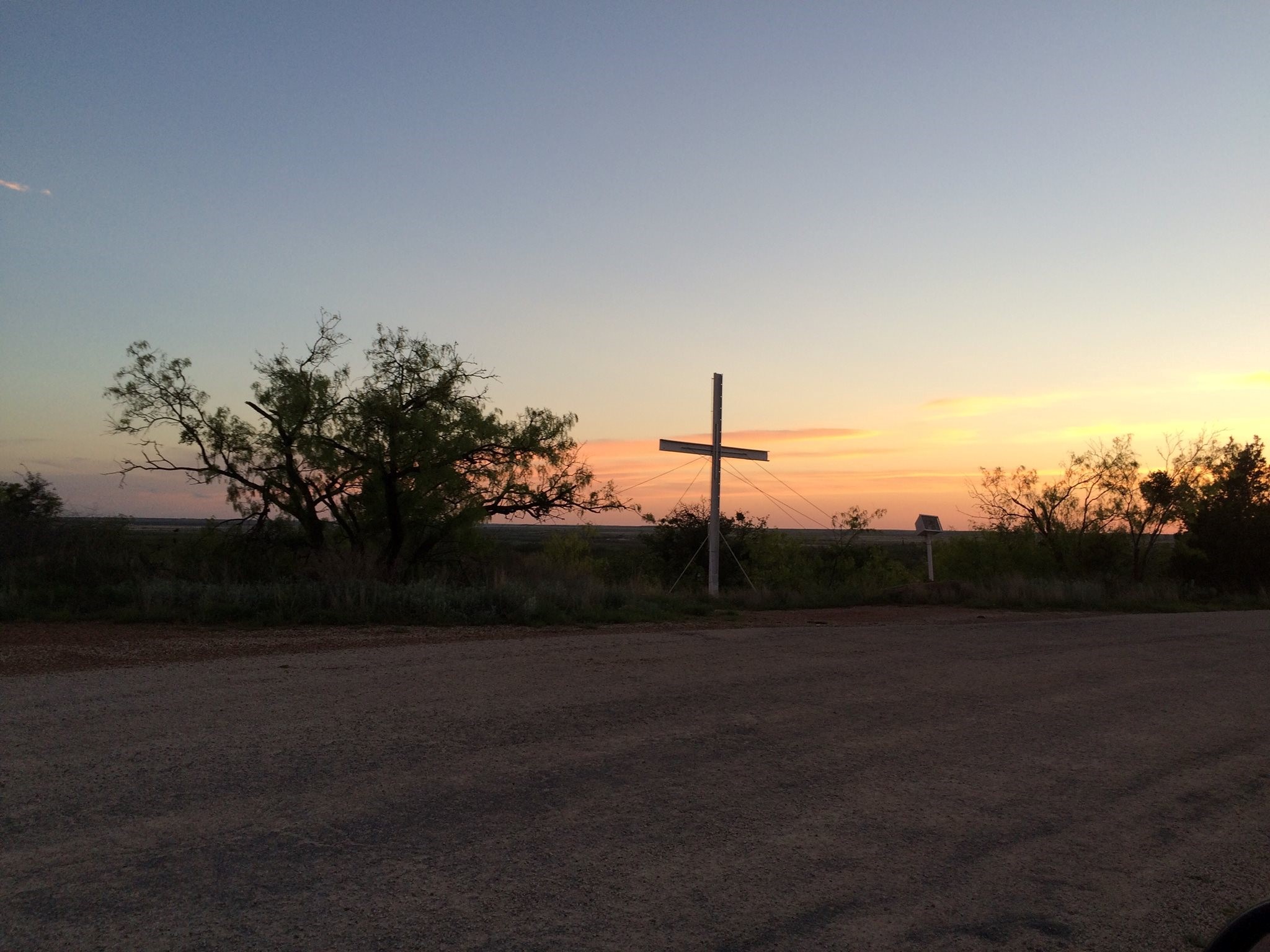 An open field with the sunset over by the mountains and a cross right on the middle of the photo.