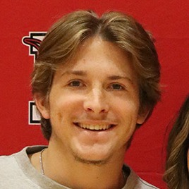 Two people stand in front of a red wall with a logo. They hold a document, smiling and posing for a picture.