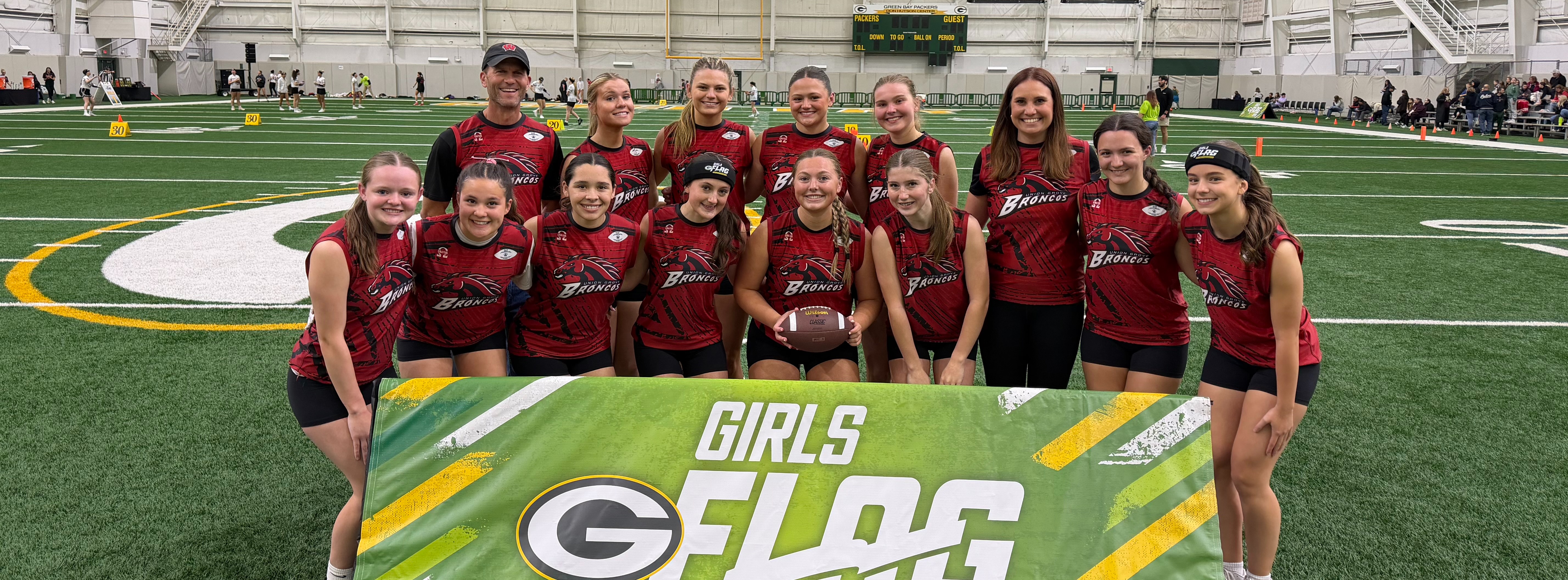 A group of women wearing red uniforms stand together on a field, holding a banner reading "Girls Flags Open."