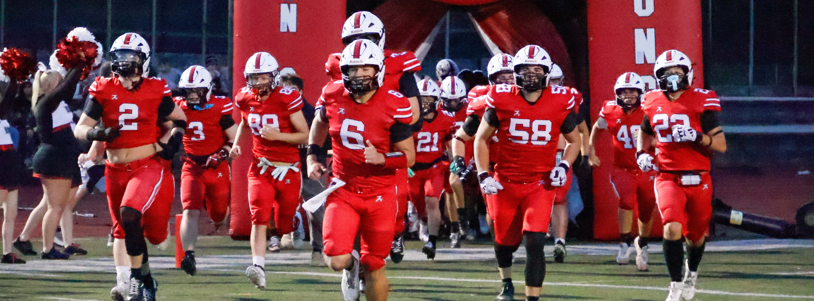 Players in red jerseys and helmets run out of a red arch on a football field.
