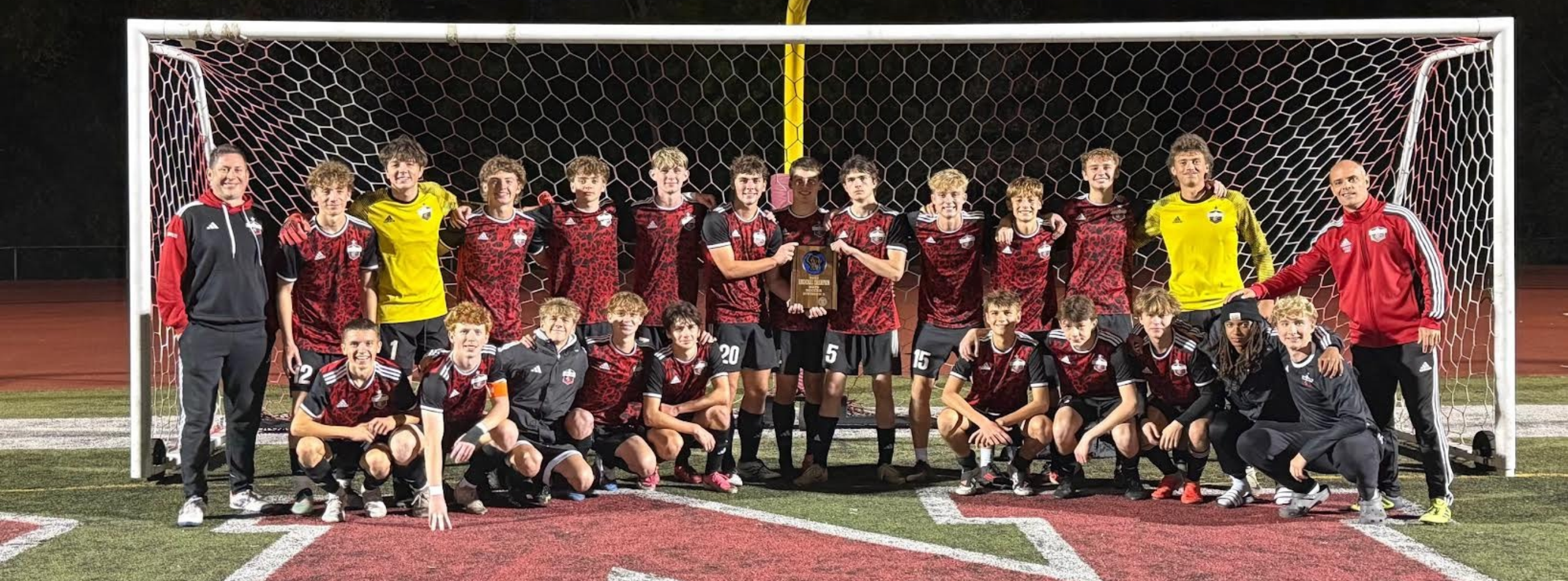 Soccer team stands in front of goal; players wear maroon jerseys, some in yellow, and black pants.
