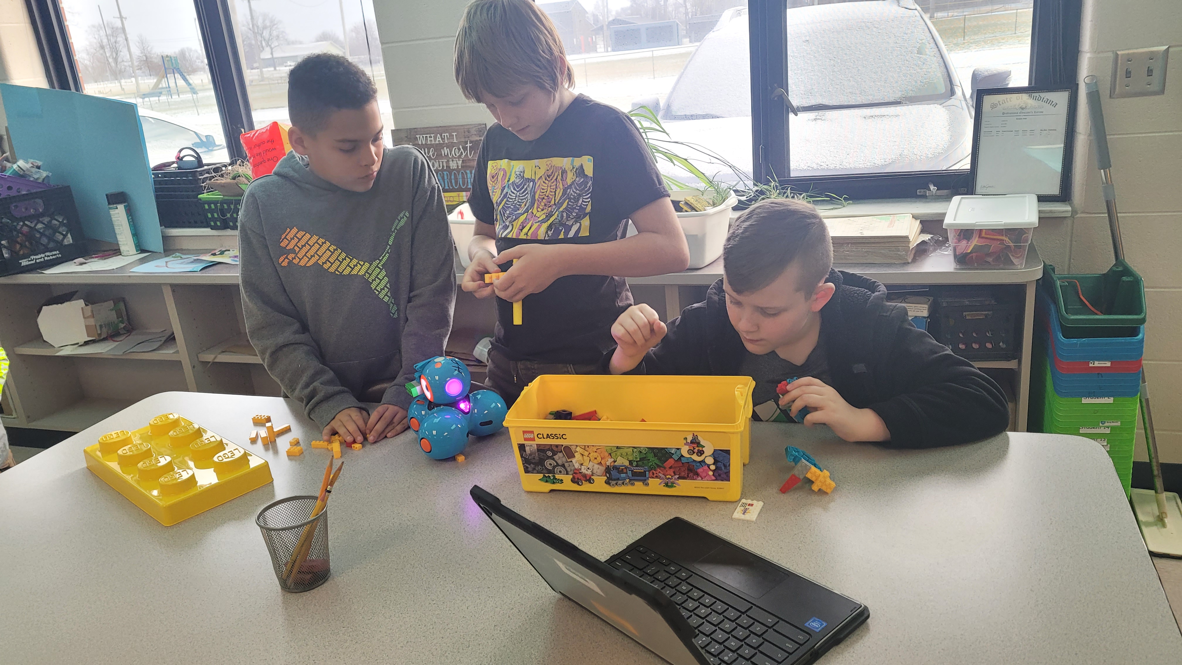 Students at a table with a coding robot, a bin of legos, and a Chromebook
