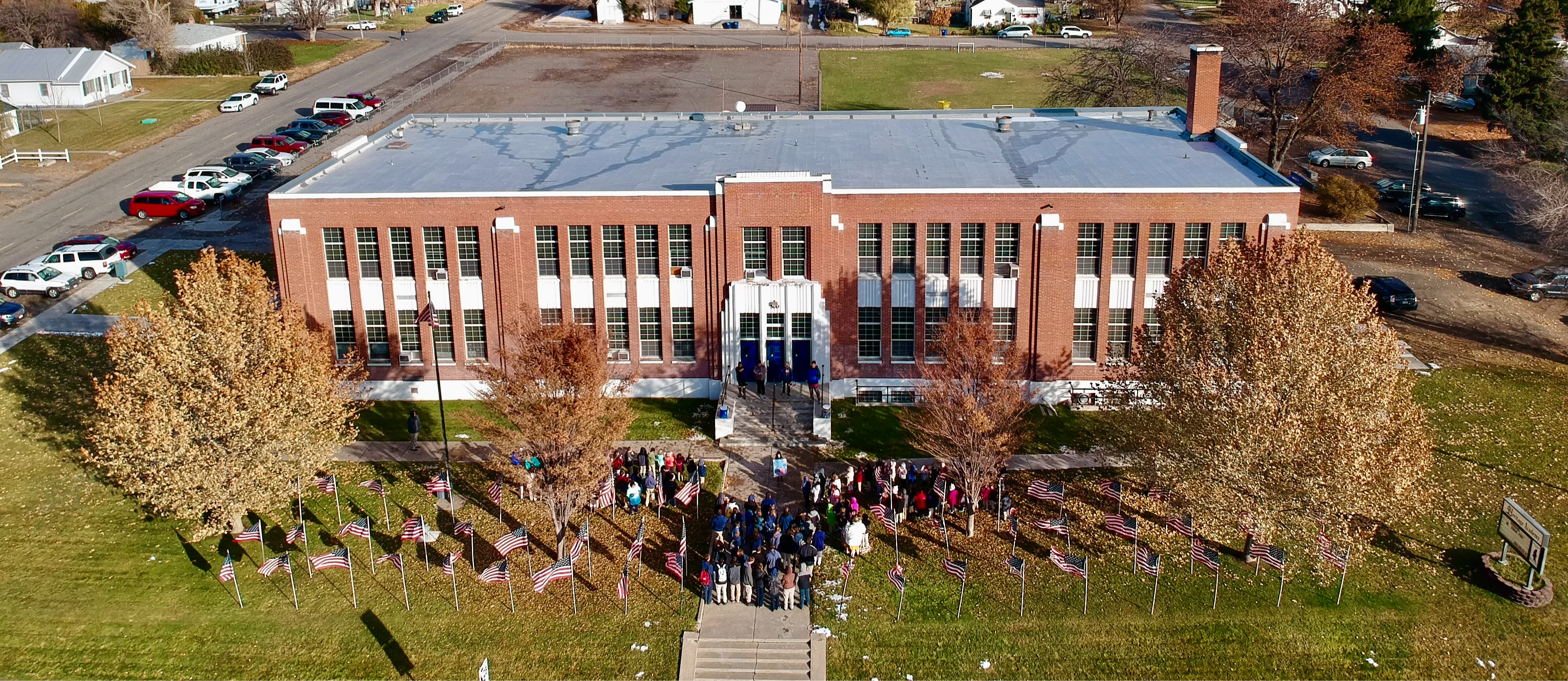 Aerial view of a large, rectangular brick building with a gray roof surrounded by trees and a lawn. People gather near a pathway lined with American flags, creating a patriotic atmosphere.