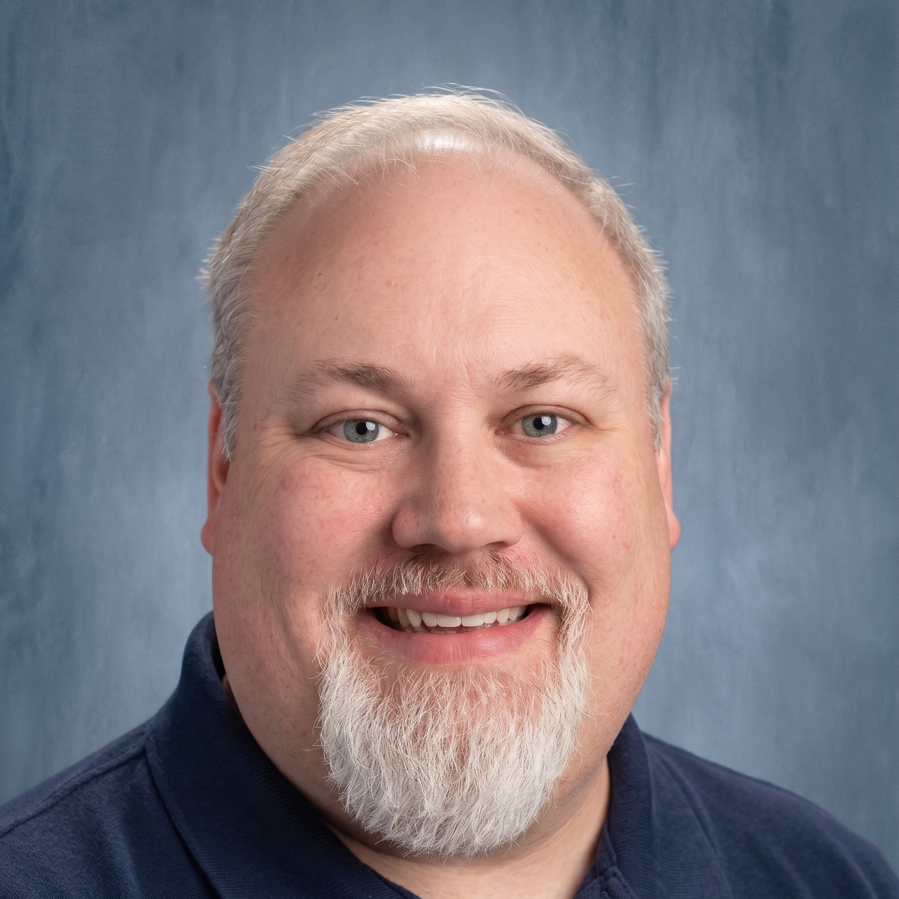 A man with a white beard and blue eyes wearing a navy blue polo shirt, smiling at the camera.