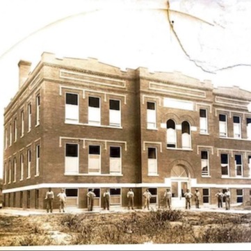Black-and-white photo of a three-story building with arched windows. A group of people stand outside, in front of the entrance.