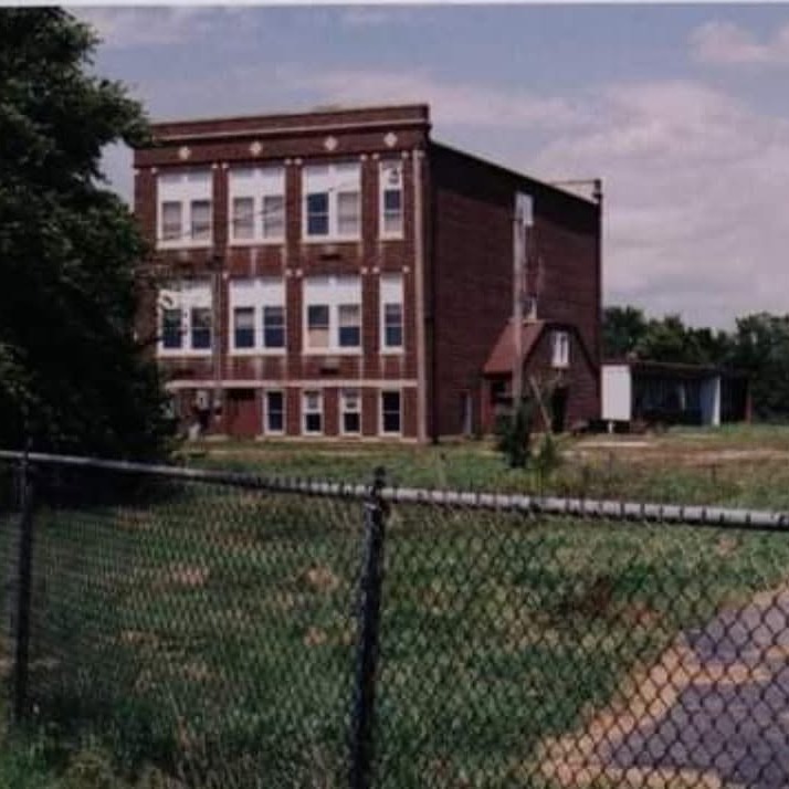 An old building with multiple windows stands in a field. A chain-link fence encloses the front. Trees and grass surround the area.