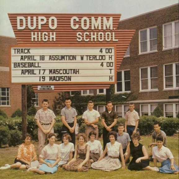 A group of people sit and stand in front of a school sign, with a building and greenery in the background.