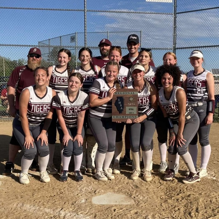 A team of women in matching uniforms stands with a trophy on a dirt field, fence behind.