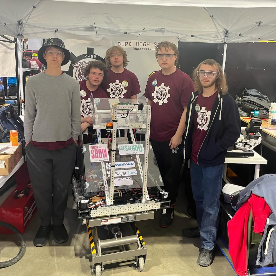 Five people in a tent, four wearing maroon shirts, stand next to a robotic device with wires.