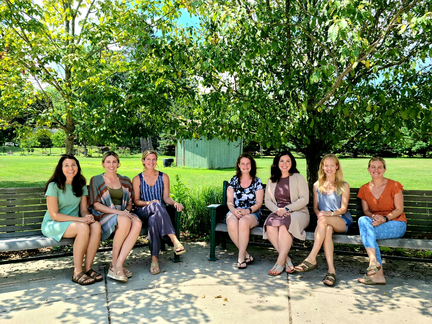 7 women sitting on a bench outside in front of trees