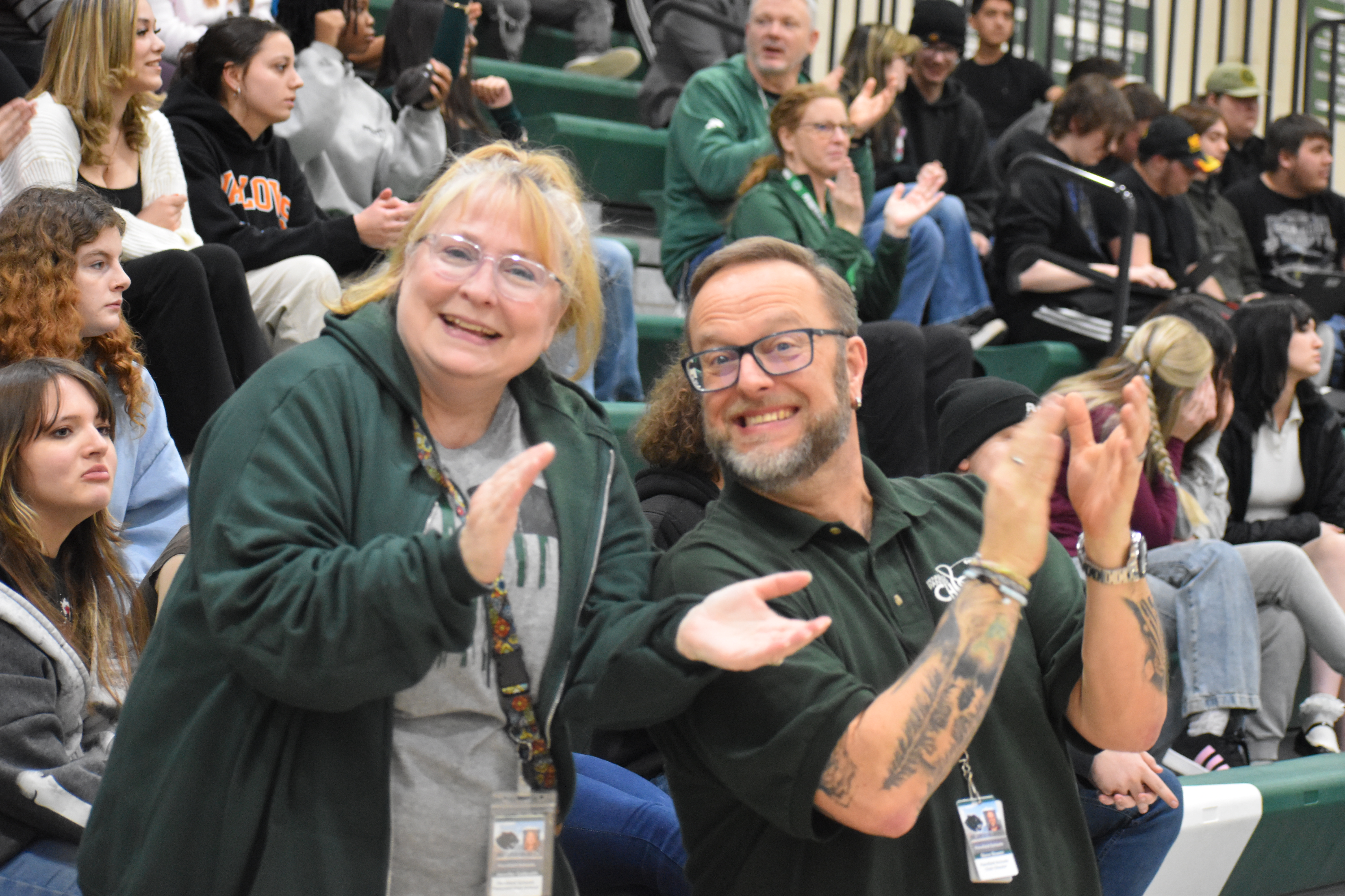 Two Pennfield Schools staff members smile and clap while seated in the gym bleachers during a school event, with students and staff seated behind them.