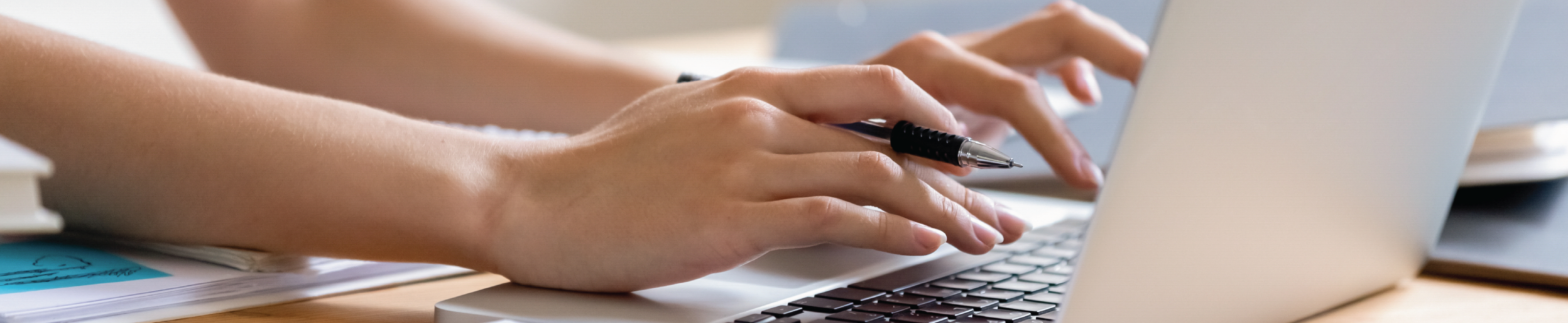 A girl holding a pen and typing on a laptop.