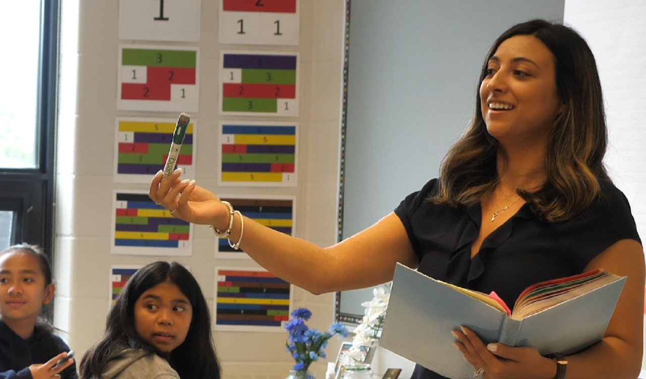 Teacher holding a marker and an open book while leading a classroom lesson, with students seated nearby.