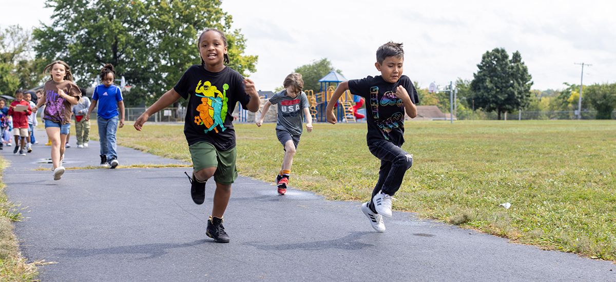 White sky above, green grass in the background, and a gray paved track with three kids running toward the camera.