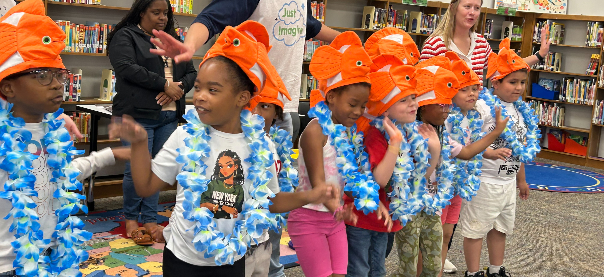 Elementary students in blue leis and goldfish hats stand in front of the library.