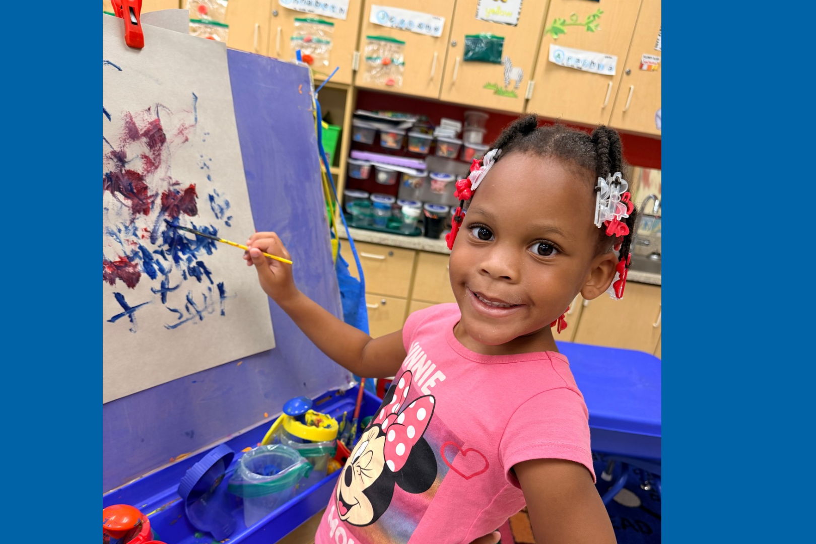 Girl holding paintbrush painting on paper in classroom. Purple board with artwork.