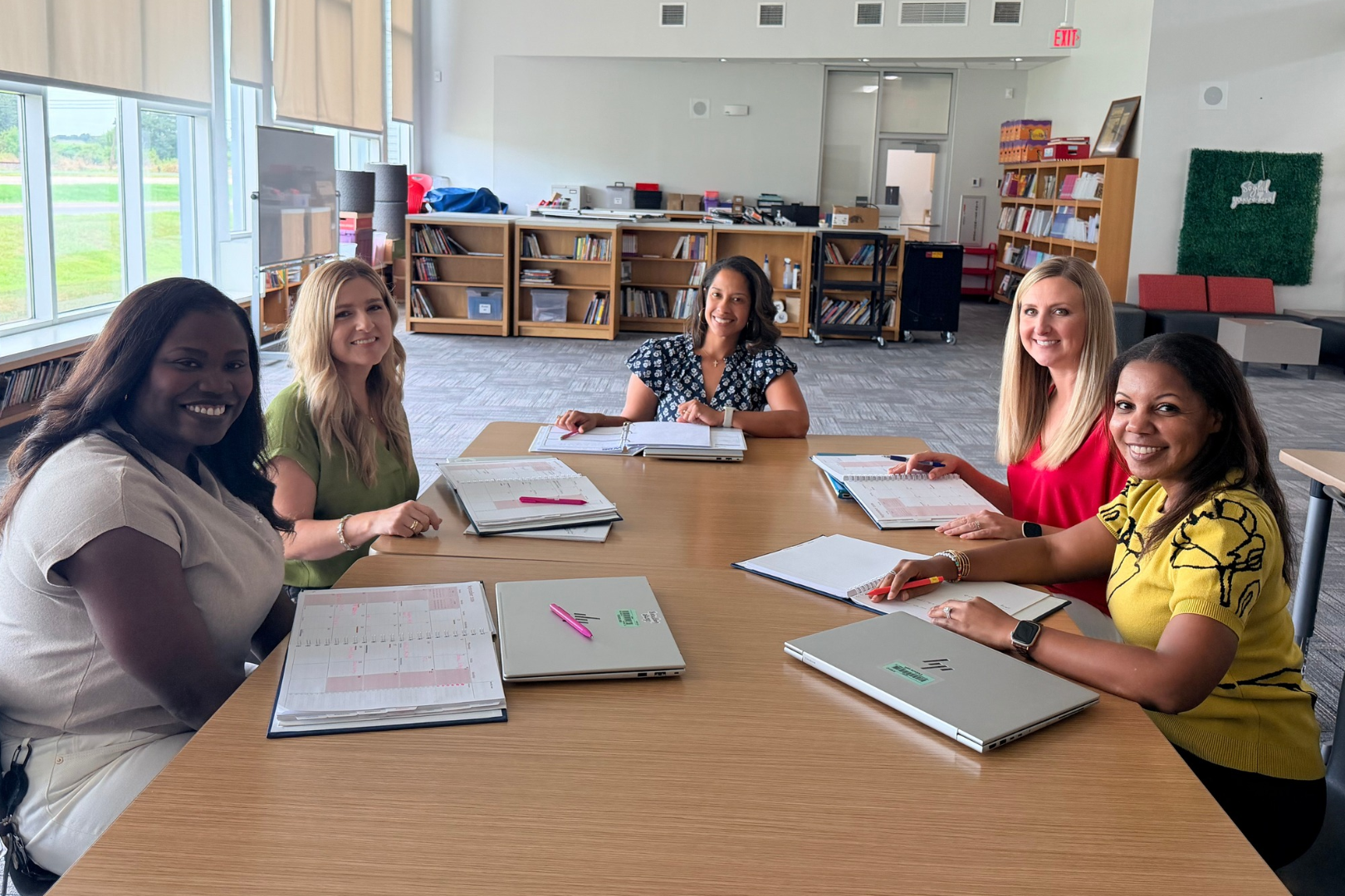 Five women sitting around a table with laptops and papers in a library with bookcases.