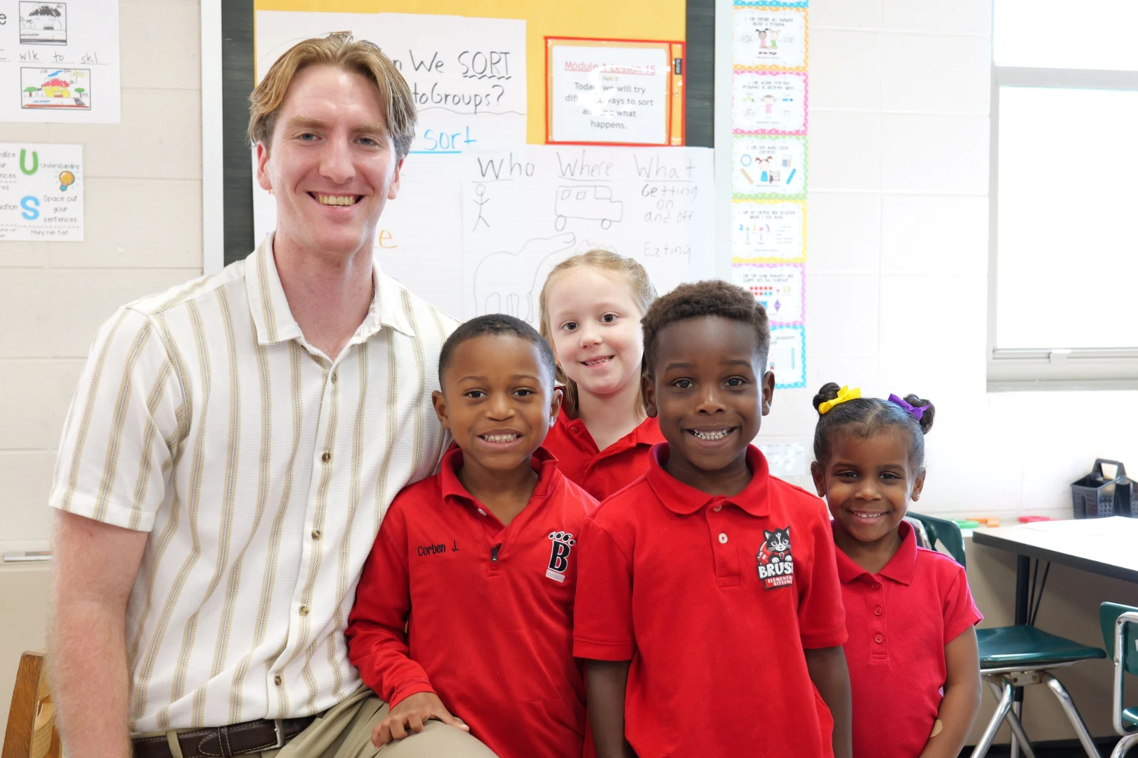 Teacher smiles with four children in a classroom. One child wears glasses, two wear name tags, and two have ponytails.