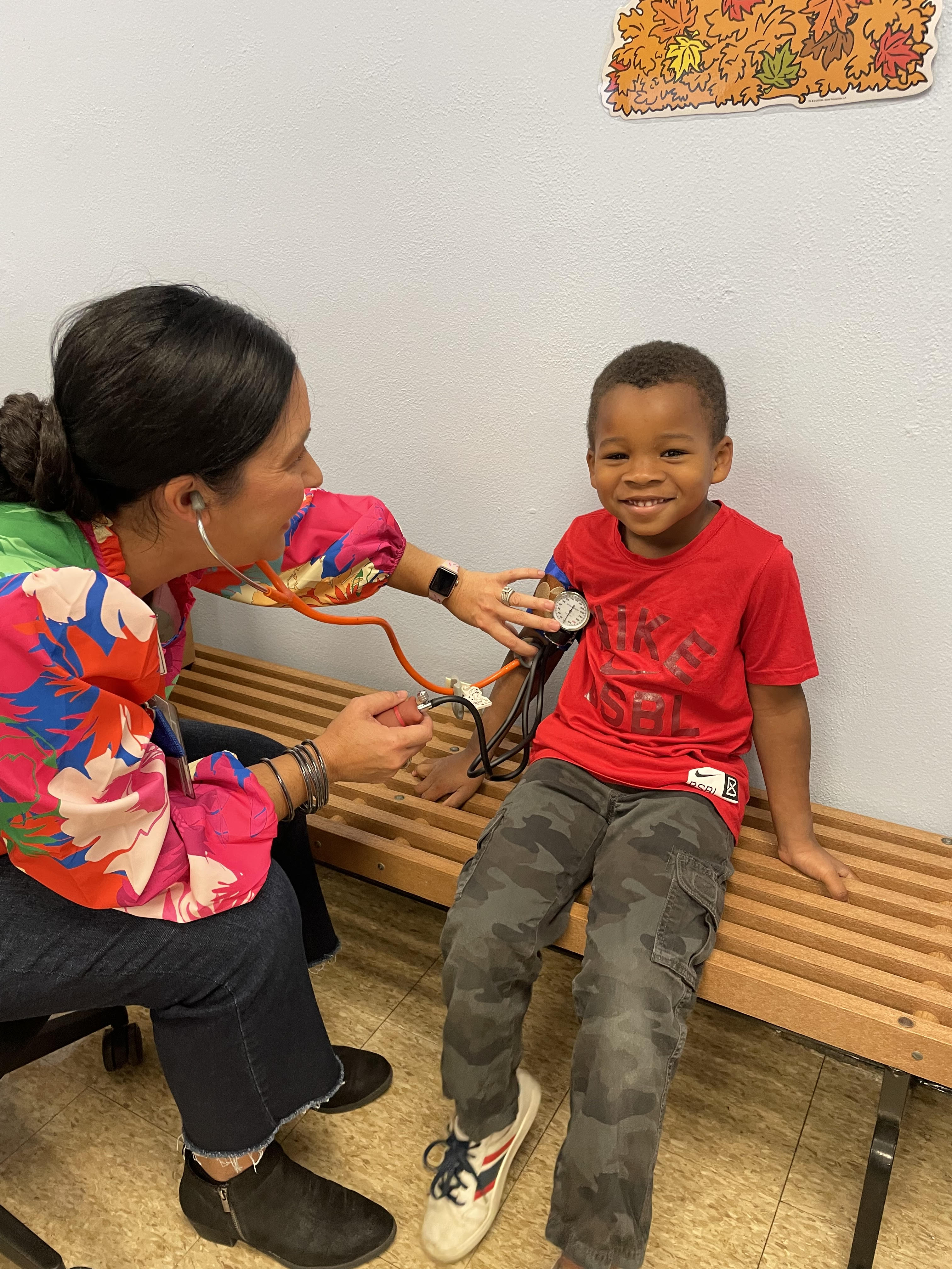 The school nurse is checking the blood pressure of a smiling student who is seated on a bench inside the school