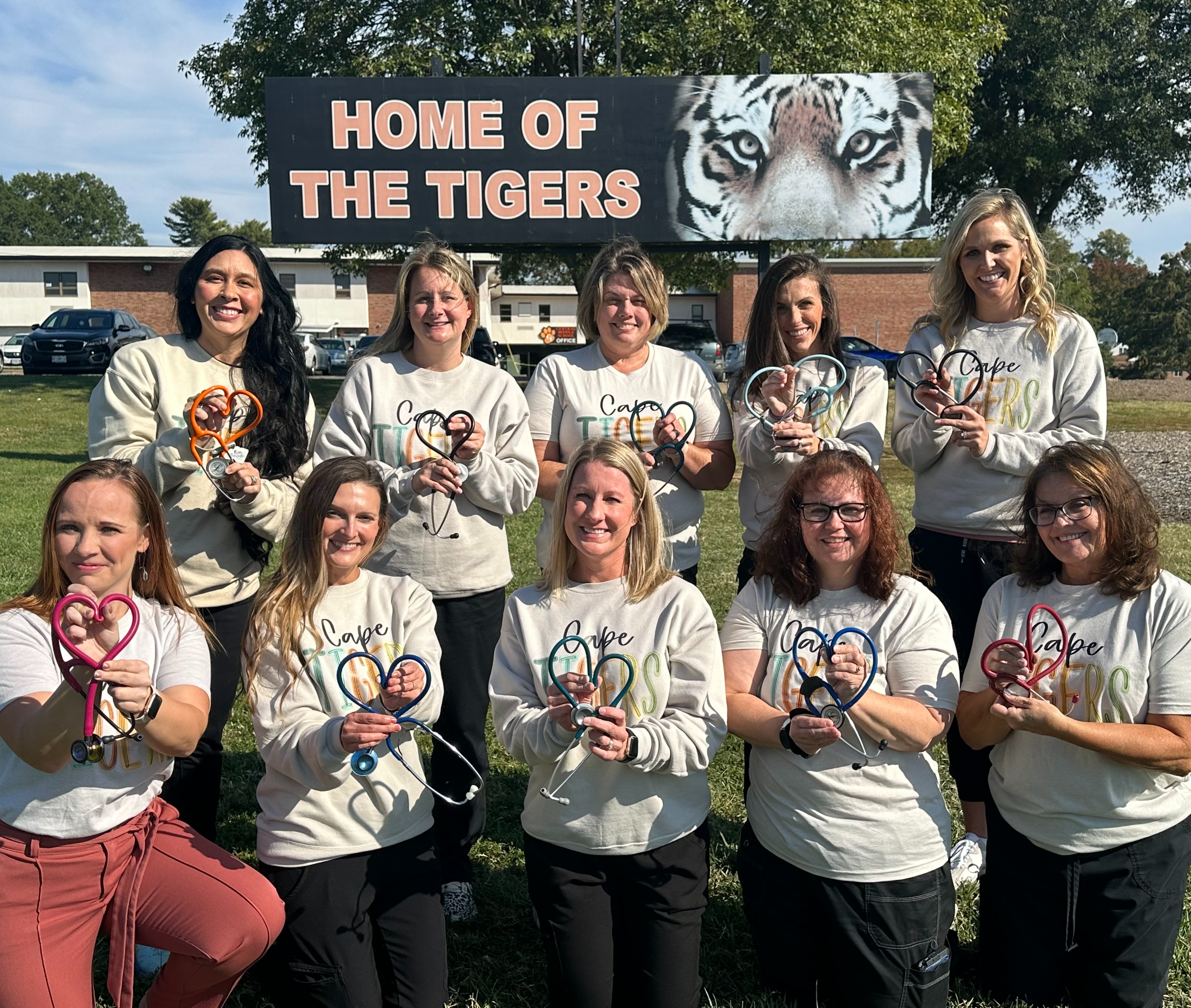 Ten school nurses standing in front of a sign that says home of the tigers, smiling for a picture 