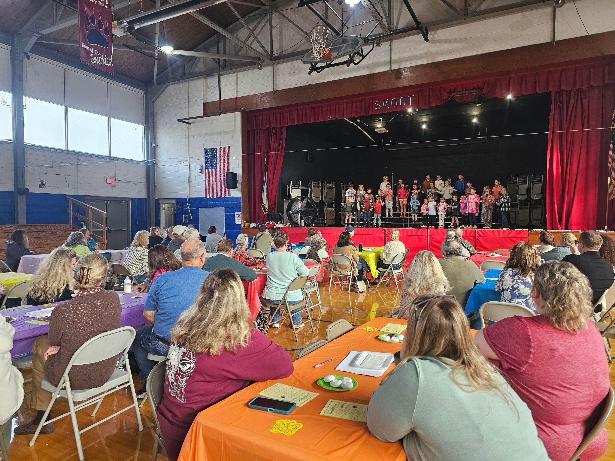grandparents day celebration in the gym with students on risers on the stage singing a song about loving them to them each at their tables. 