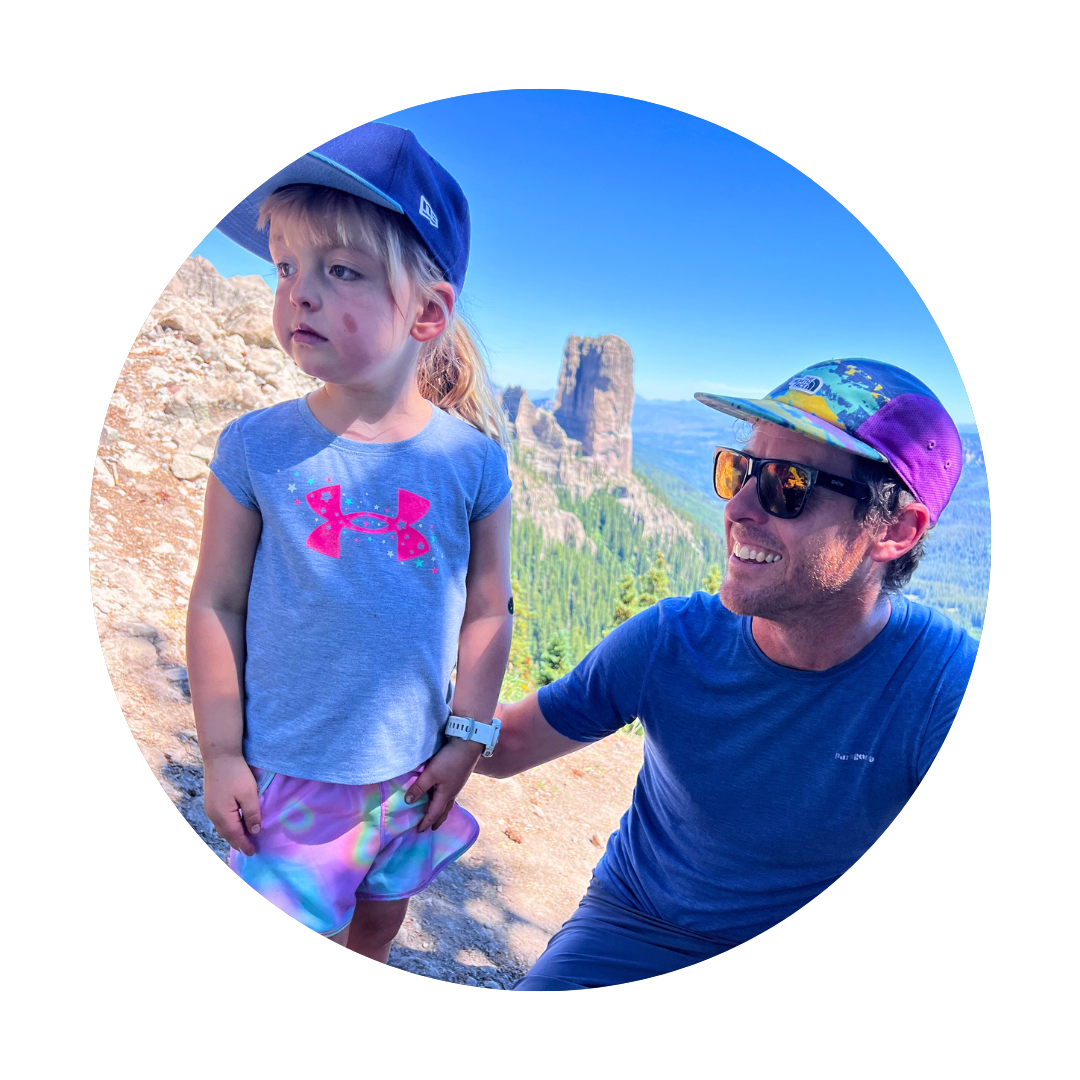 A man and a young child, both wearing caps, enjoy a scenic outdoor adventure with a forested landscape and rocky formations in the background.