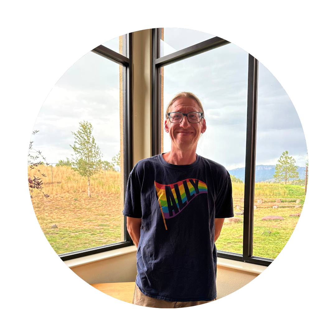 A man standing indoors with large windows showing a scenic outdoor view of trees and fields.