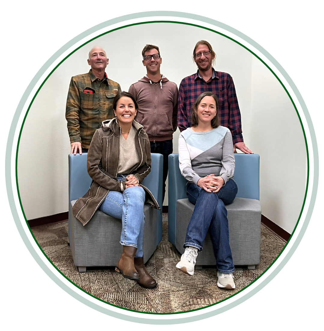 School Board Member Group portrait of five adults indoors. Two people are seated on square chairs in the front, smiling, with three people standing behind them against a plain wall.