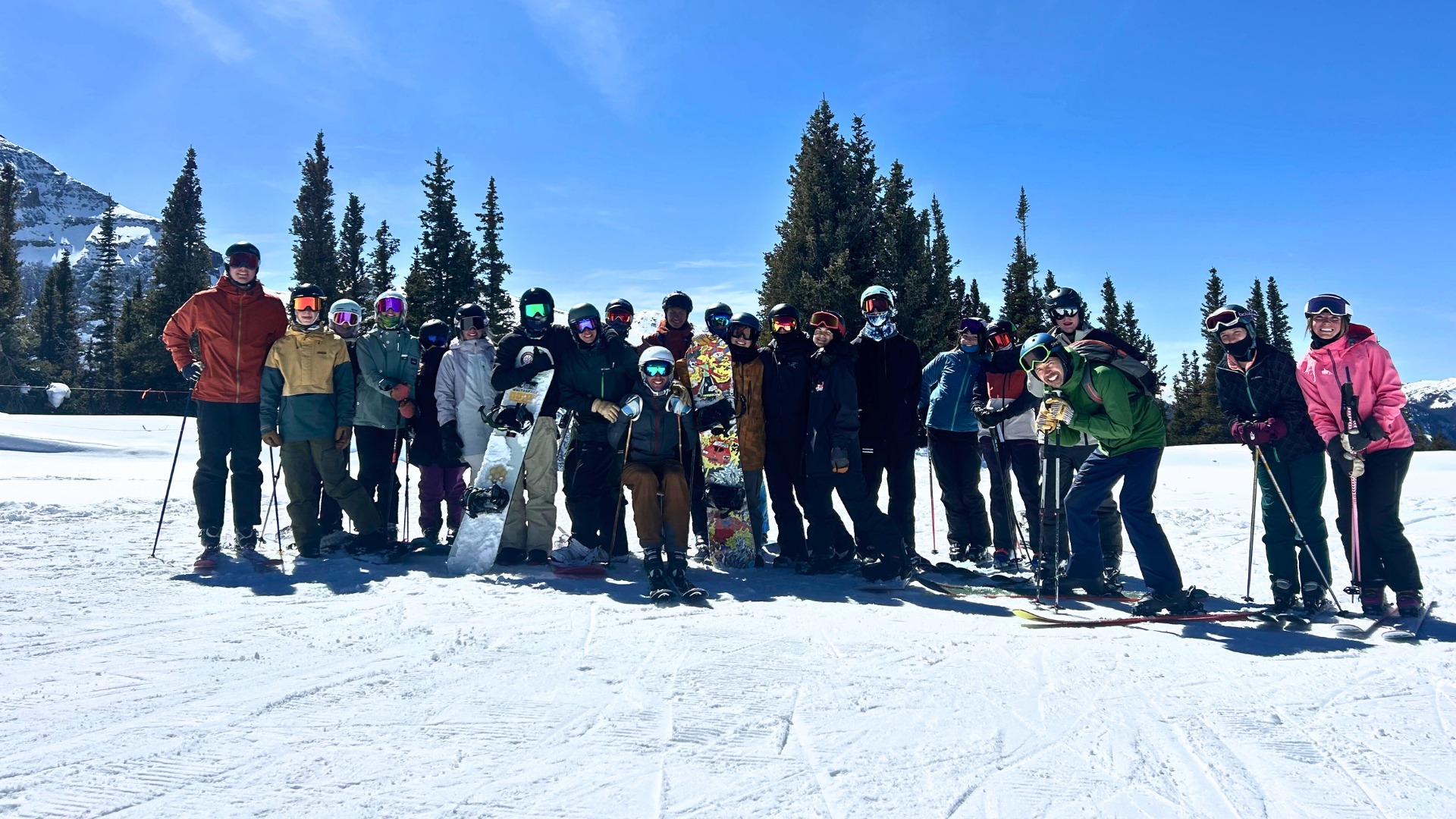 Group of students and adults in ski gear posing together on a snowy mountain under a clear blue sky.