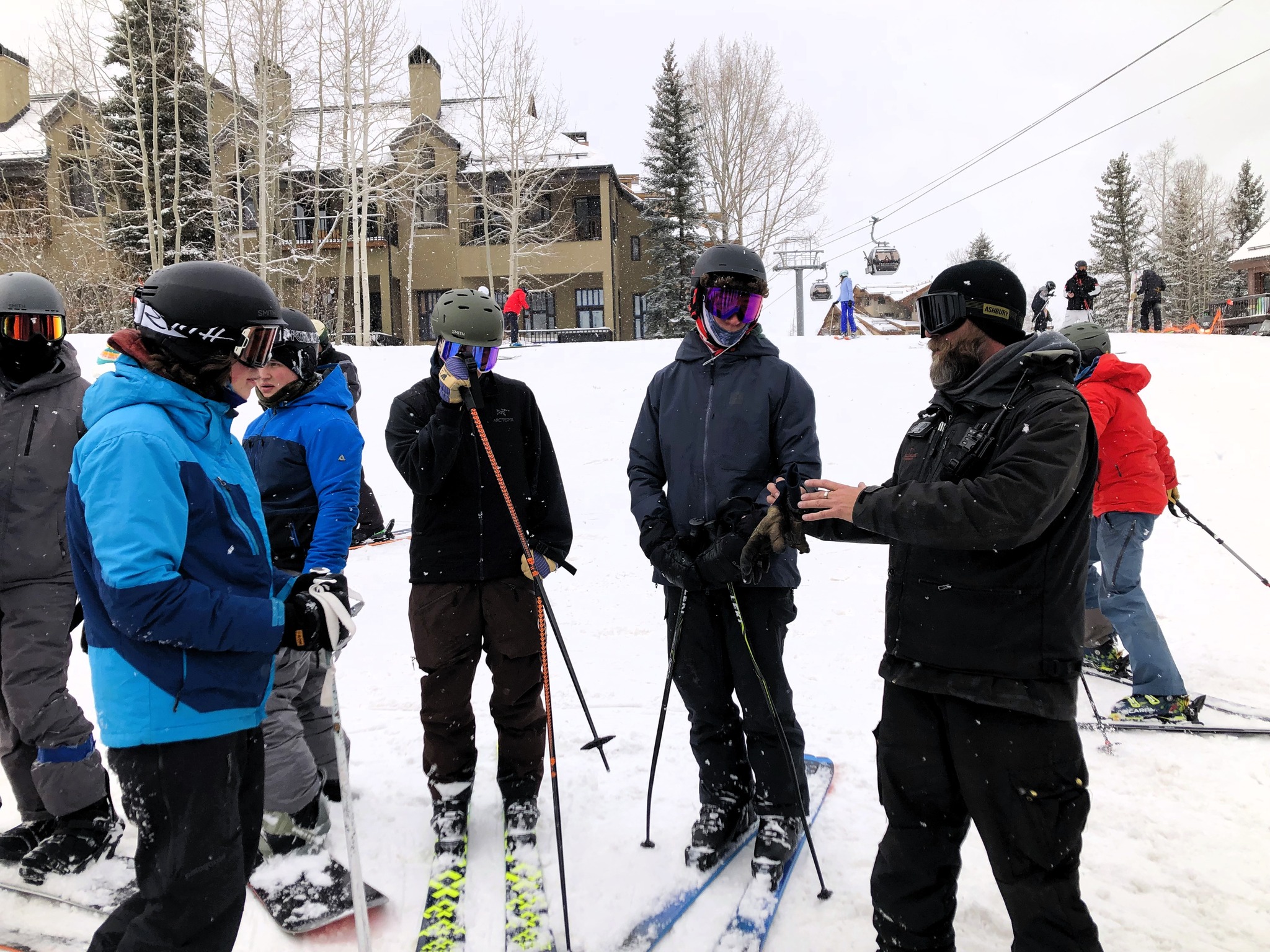 Students in ski gear stand on a snowy slope while an instructor gives directions.