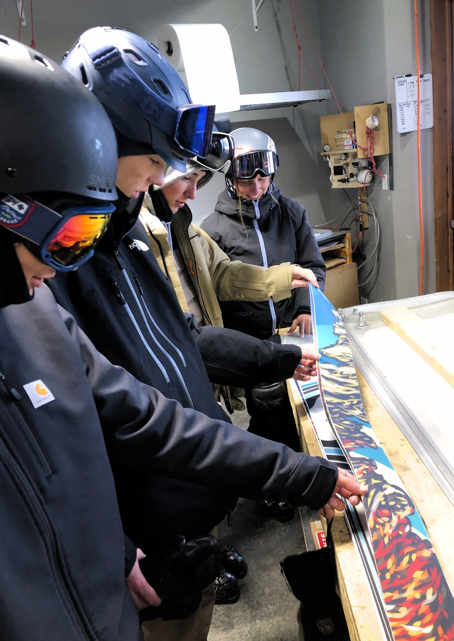 Students in ski gear examine and tune a ski on a workbench indoors.
