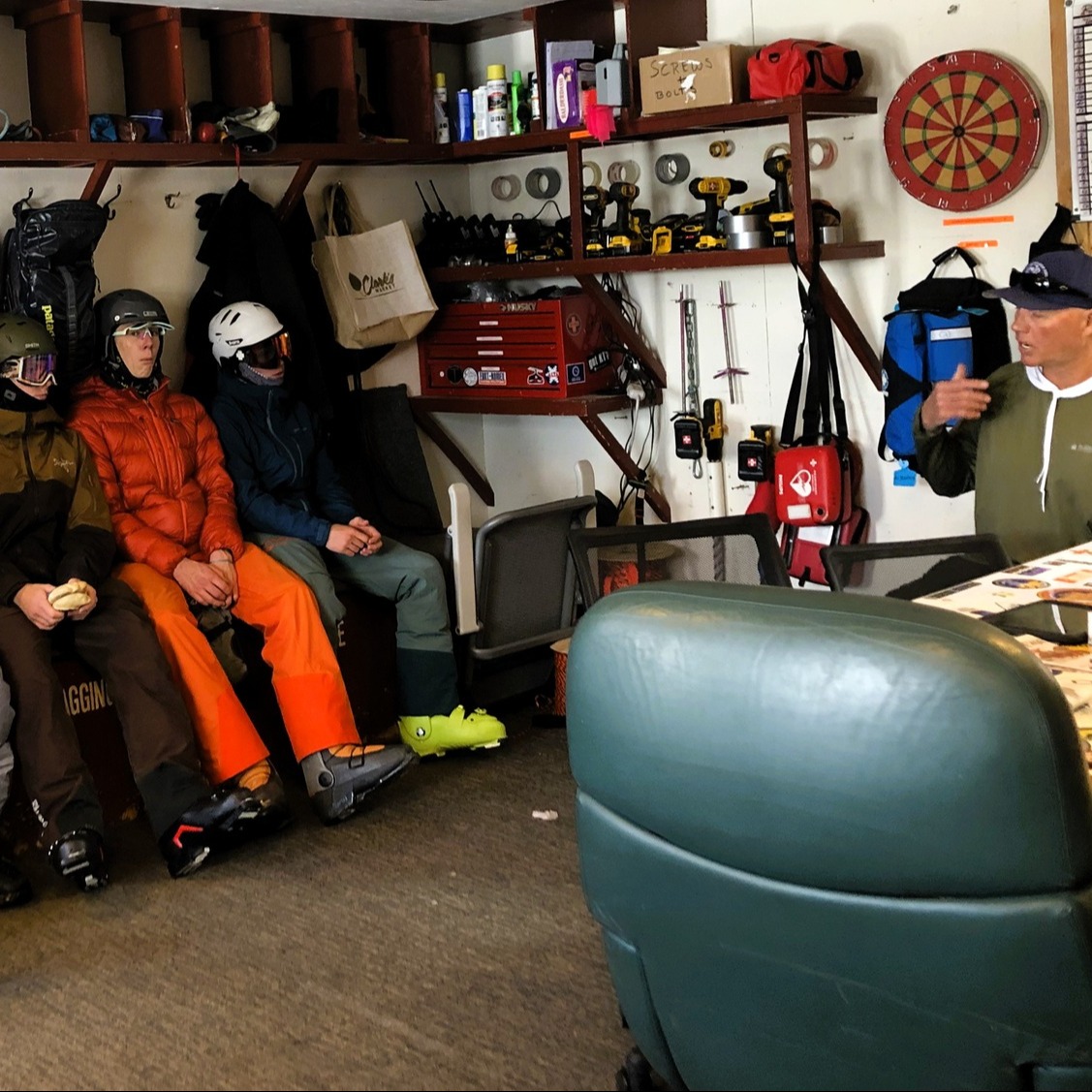Students in ski gear sit in a ski patrol room listening to a safety briefing.