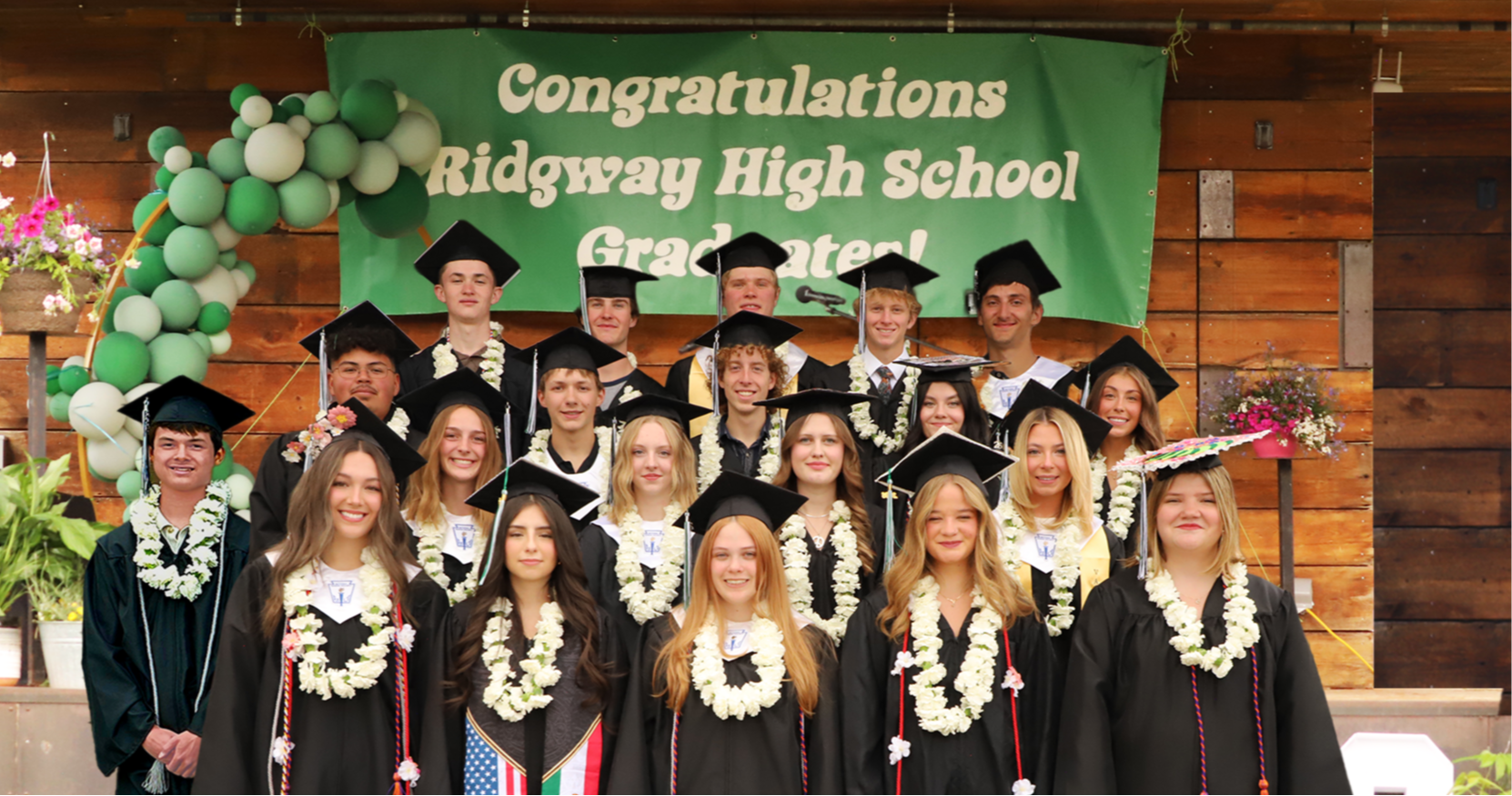 Group photo of Ridgway High School’s Class of 2026 graduates in caps and gowns standing in front of a green congratulatory banner at graduation.