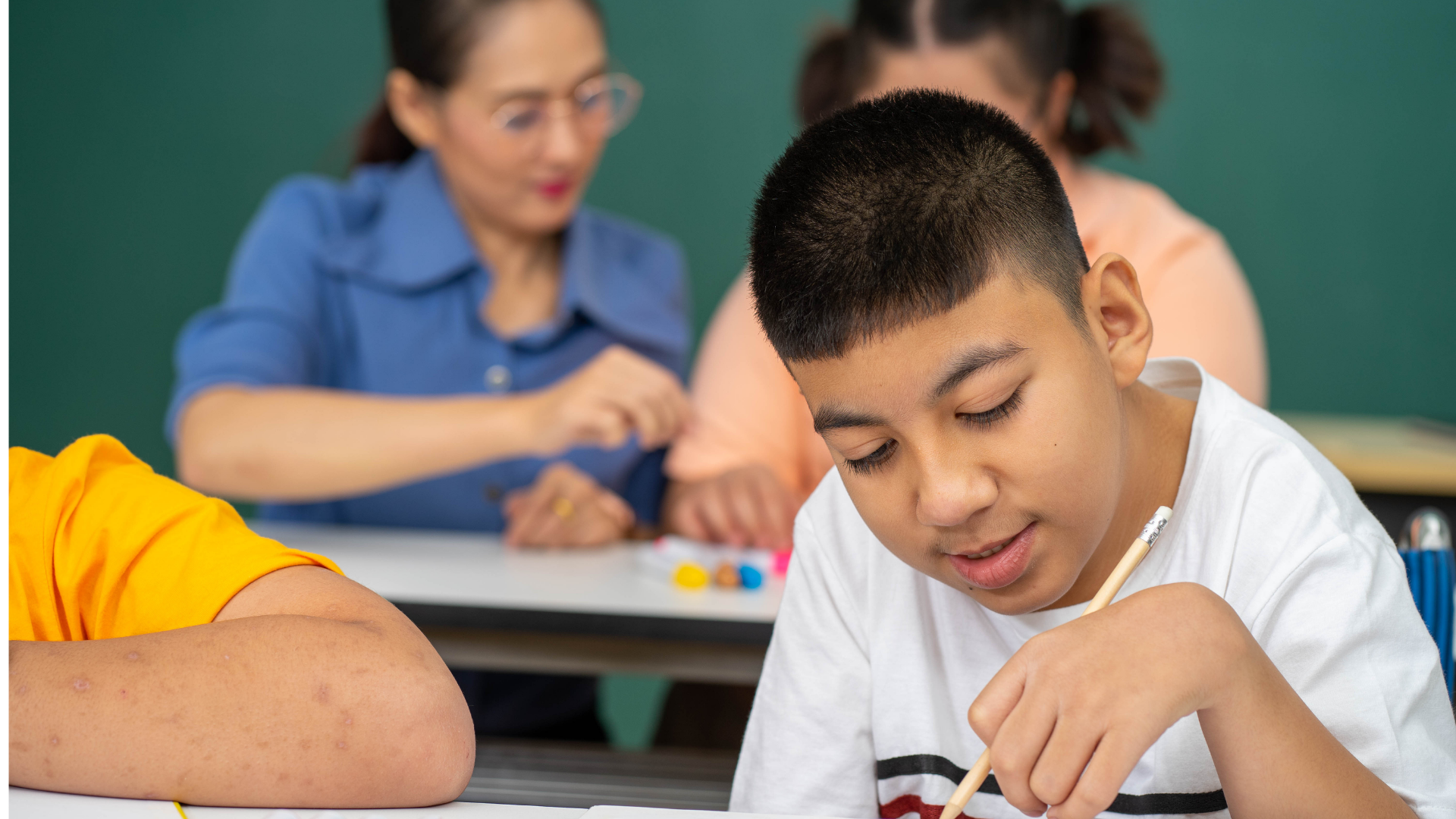Photo of a hispanic adolescent male student writing at table with teacher and other students in background.