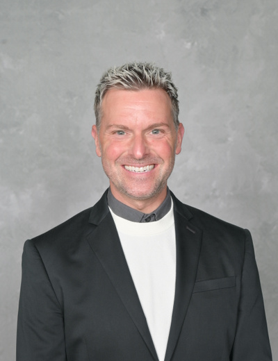 A man with gray hair wearing a black suit with a white shirt underneath smiles at the camera.
