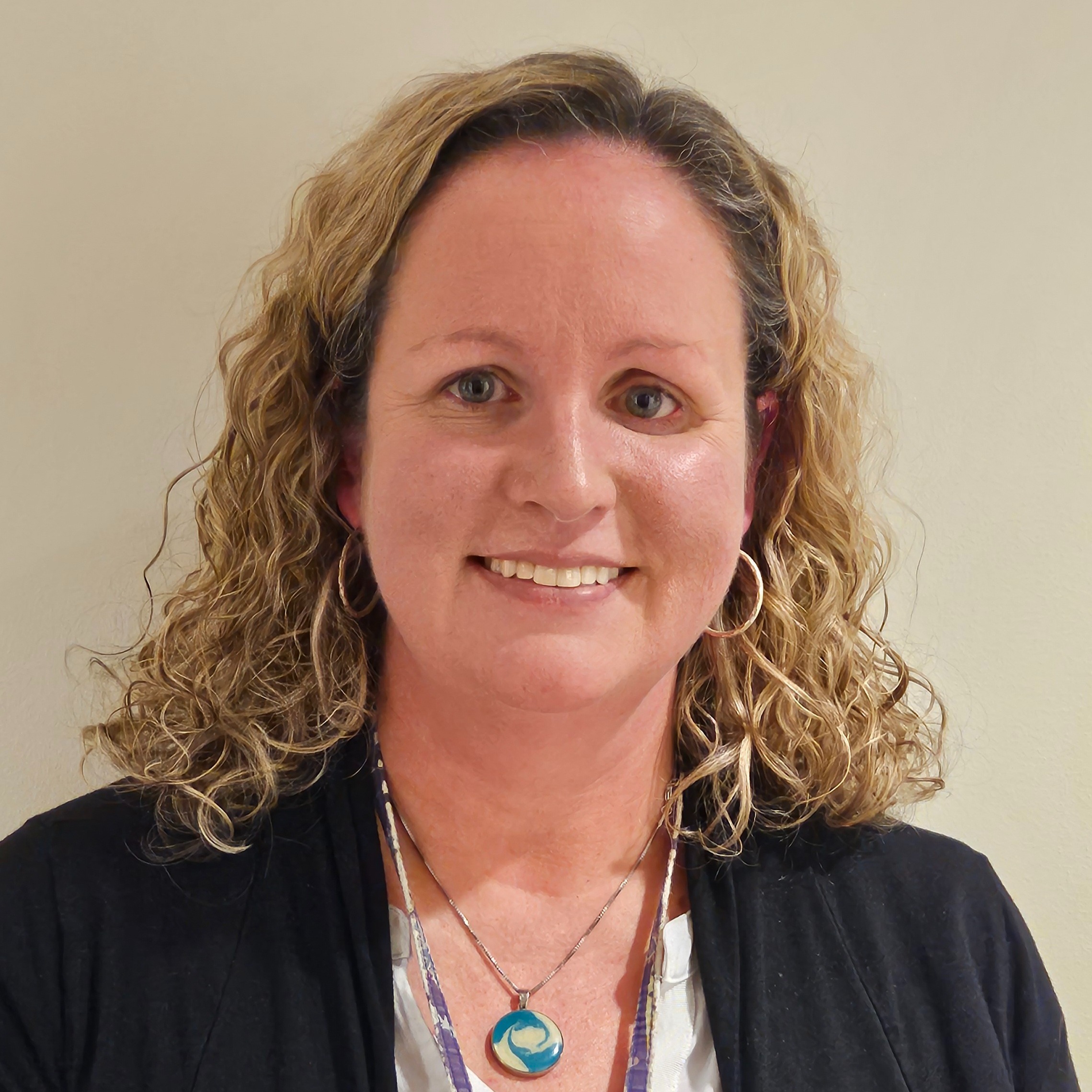 Woman with curly blonde hair smiling at the camera. She wears a white shirt and black cardigan, along with a necklace.