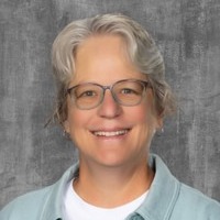 A woman with short gray hair and glasses smiles. She wears a white shirt and light blue shirt. Gray textured background.