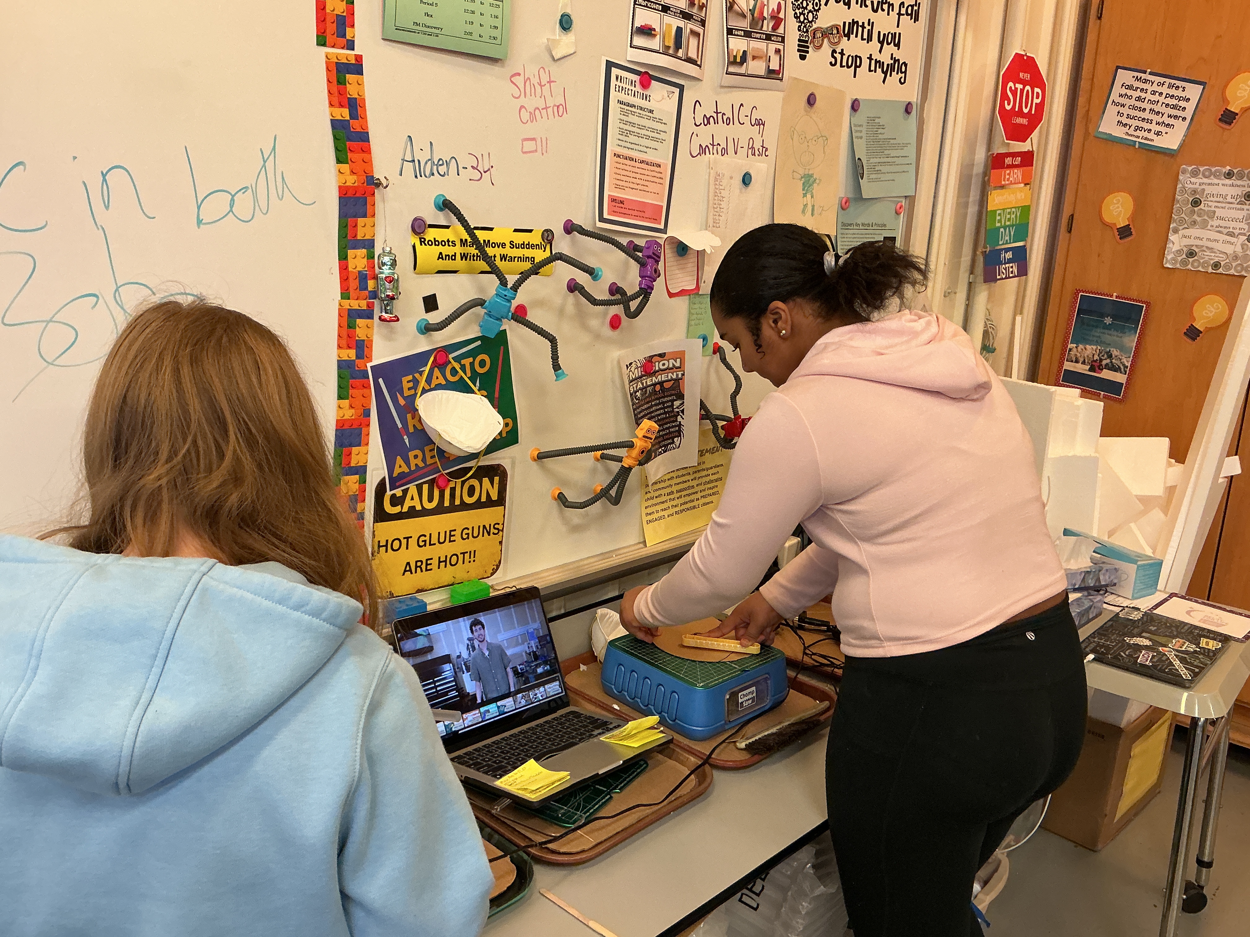 Two people in a classroom, one using a laptop, the other in a pink hoodie, at a desk.