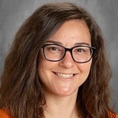 Woman with brown hair, wearing glasses, smiling, and standing against a gray background.