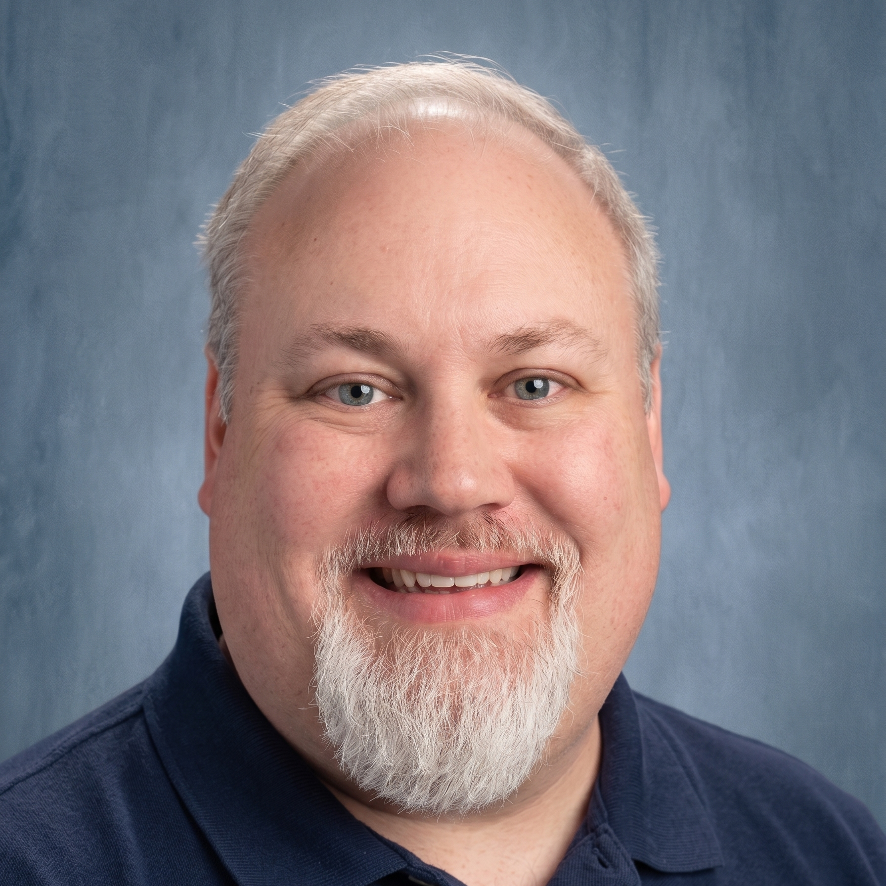 A smiling man with white hair and a white beard, wearing a navy polo shirt, in front of a blue background.