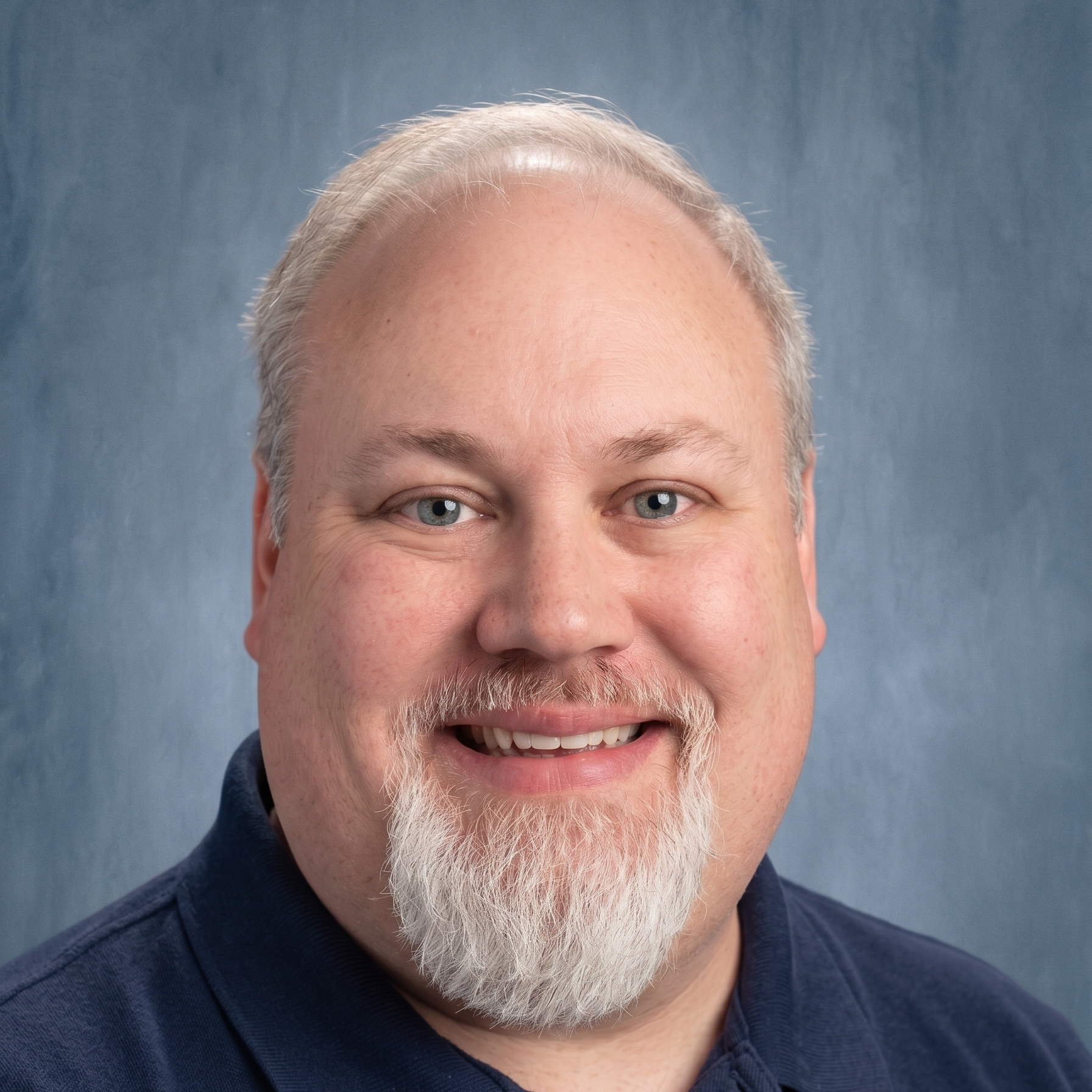 A man with a white beard and blue eyes smiles for a photo. He wears a navy blue polo shirt against a blue background.
