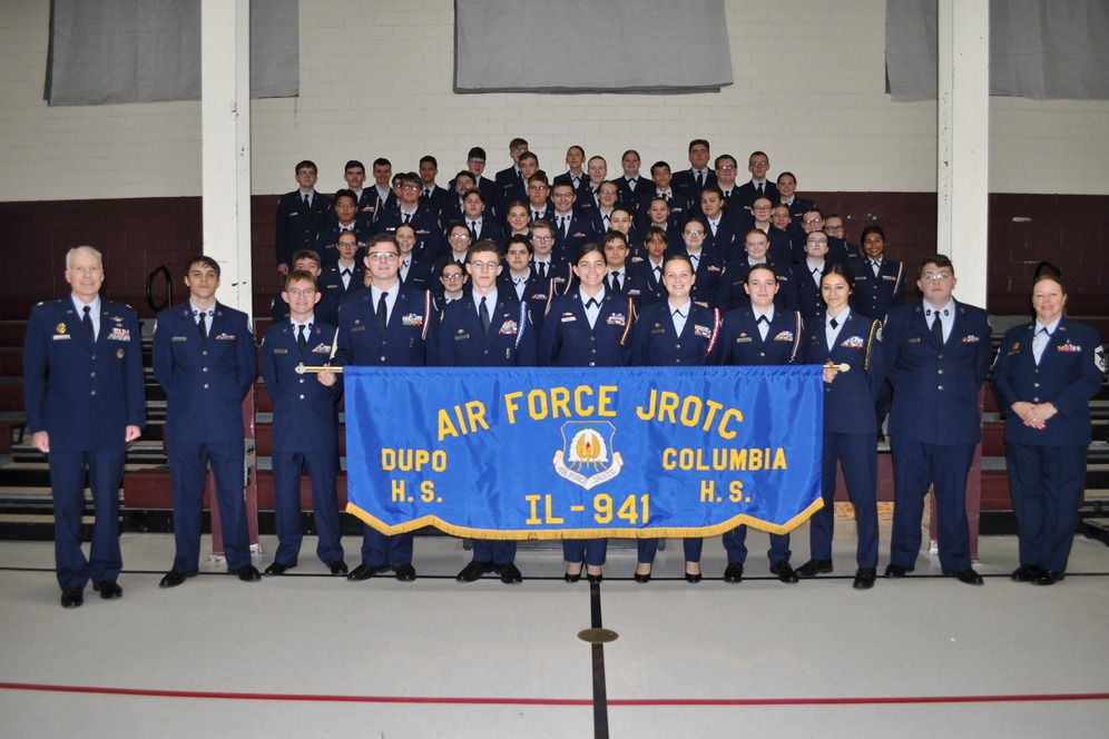 A group of uniformed individuals, possibly cadets, standing together. A blue banner with white writing is held by some.