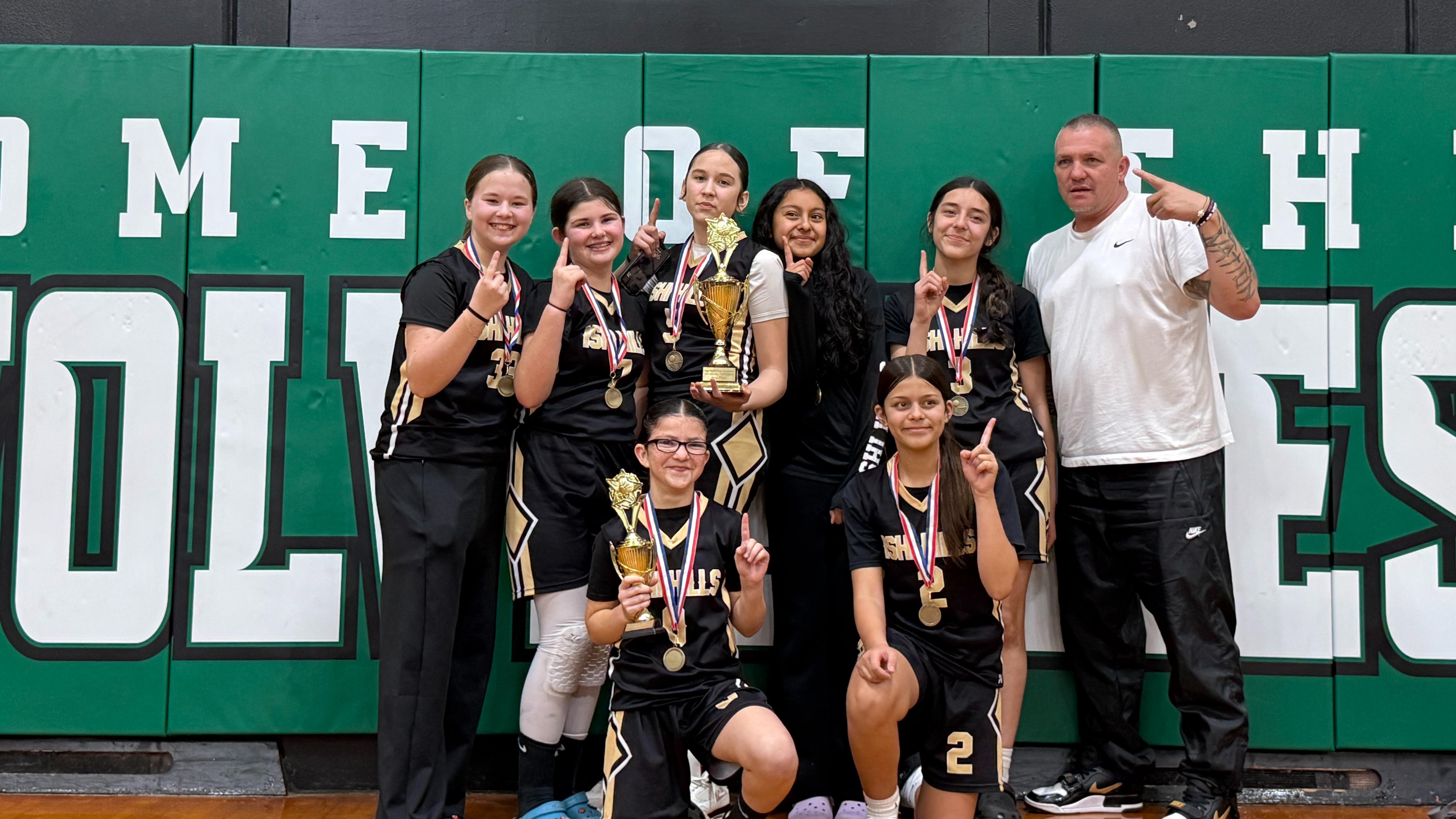 Group of basketball players holding medals, posing in front of a green wall with "HOME SCHOOL" written on it.