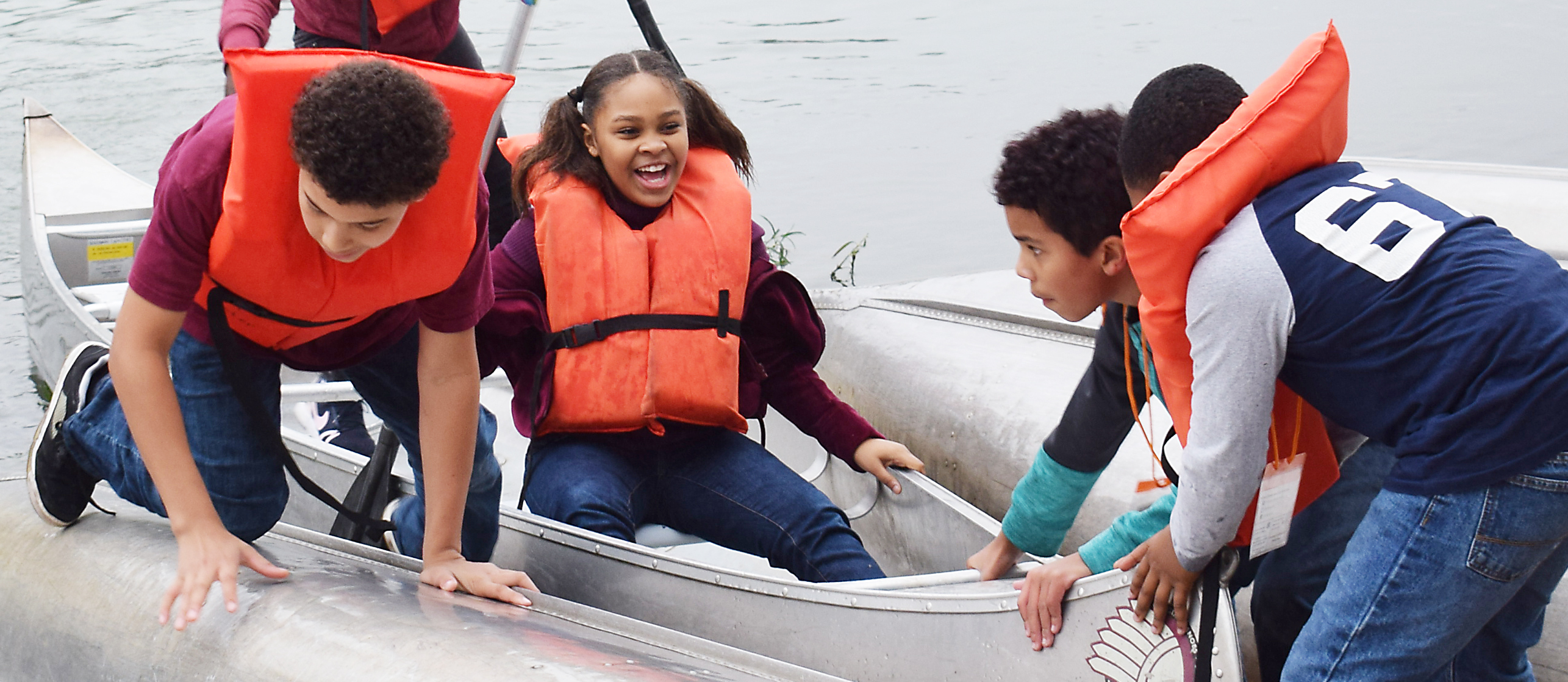 Students wearing life jackets laugh as they push a canoe into water