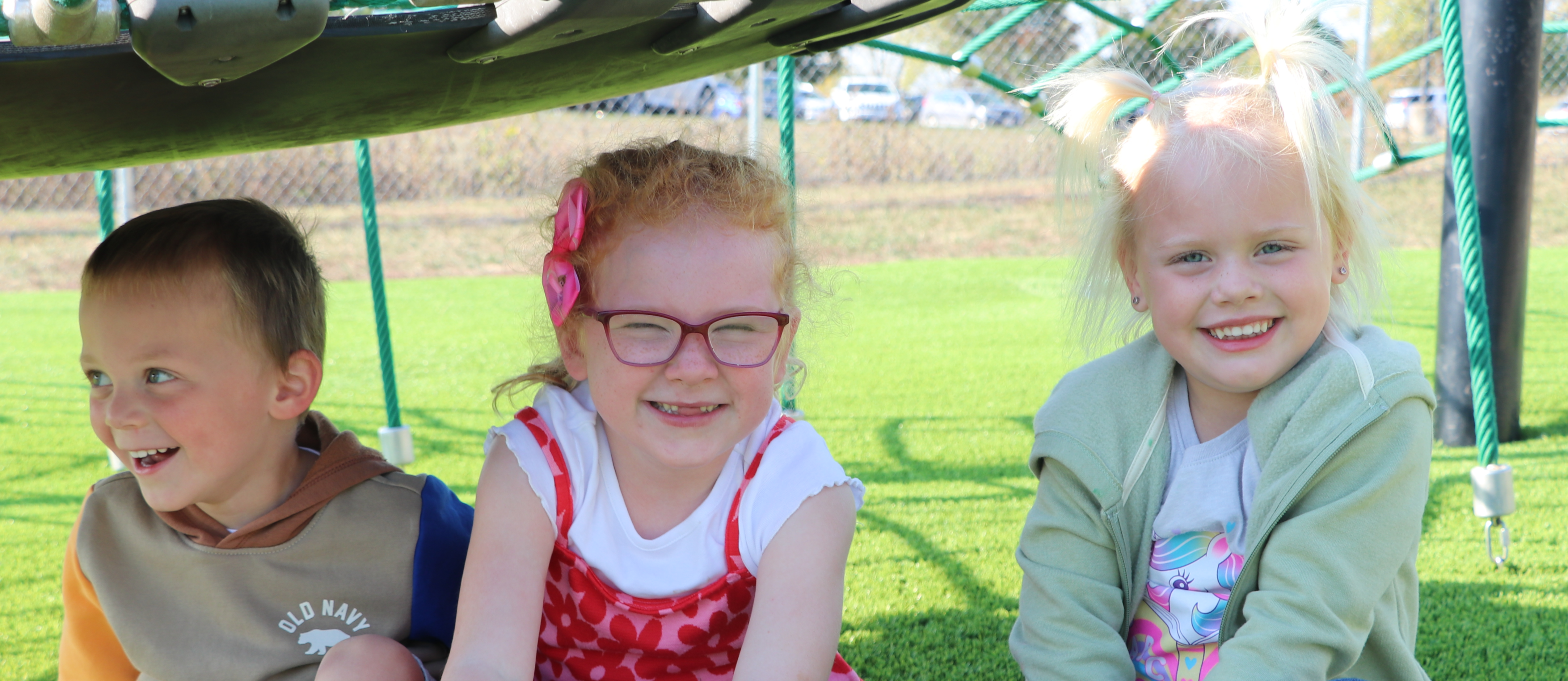 Three Warrior Preschool students smile as they sit under playground equipment.