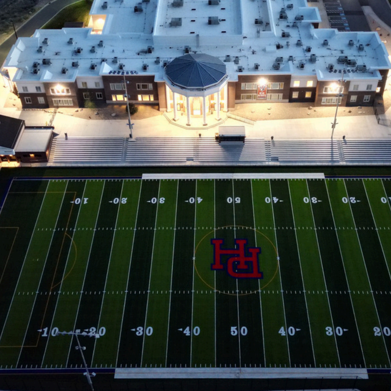Aerial view of a football field with a large building in the background. The field has markings and a logo.