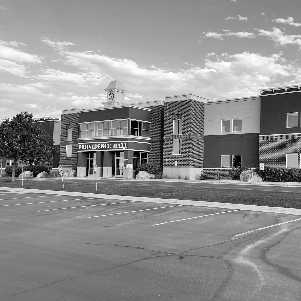 A monochromatic image of a large building with a tree in front and a large parking lot.