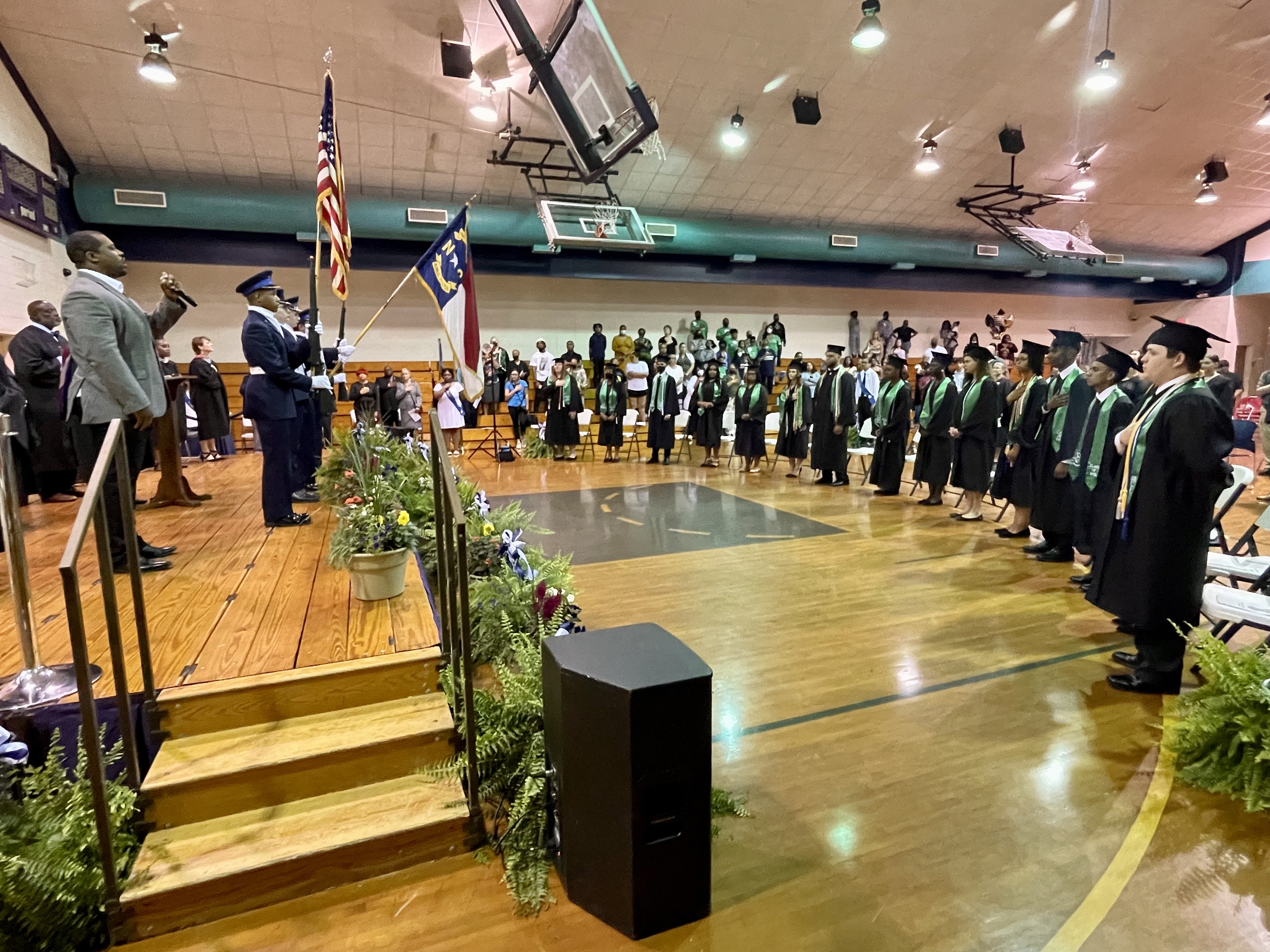 Honor guard presenting american and nc flags on stage at high school graduation, with graduates watching