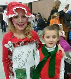a young girl in a red hat and glasses dressed as Mrs. Claus and a younger boy in a red shirt and green scarf