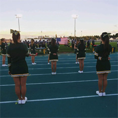 cheerleaders standing in formation on a football field and facing the American flag at dusk